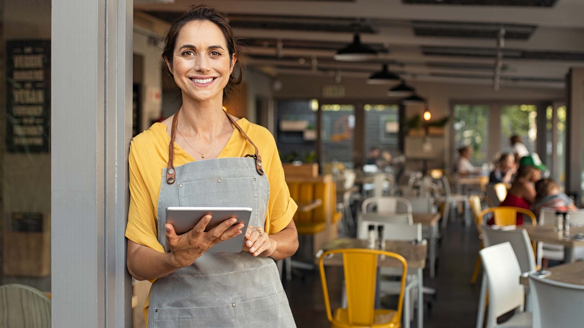 A smiling woman wearing a yellow shirt and gray apron holds a tablet while standing in a modern café or restaurant, with customers and yellow chairs visible in the background.
