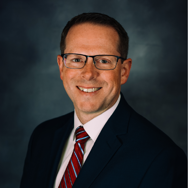 Professional headshot of a smiling man wearing glasses, a dark blazer, white dress shirt, and red striped tie against a dark blue background.