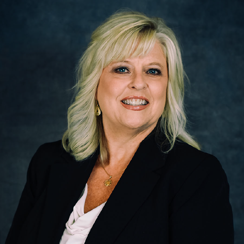 Professional headshot of a woman with blonde layered hair, wearing a black blazer over a white blouse, gold hoop earrings, and a pendant necklace, smiling at the camera against a dark blue background.