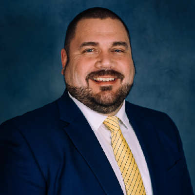 Mortgage lending team - Professional headshot of a man wearing a dark blue blazer, white dress shirt, and yellow striped tie against a blue background.