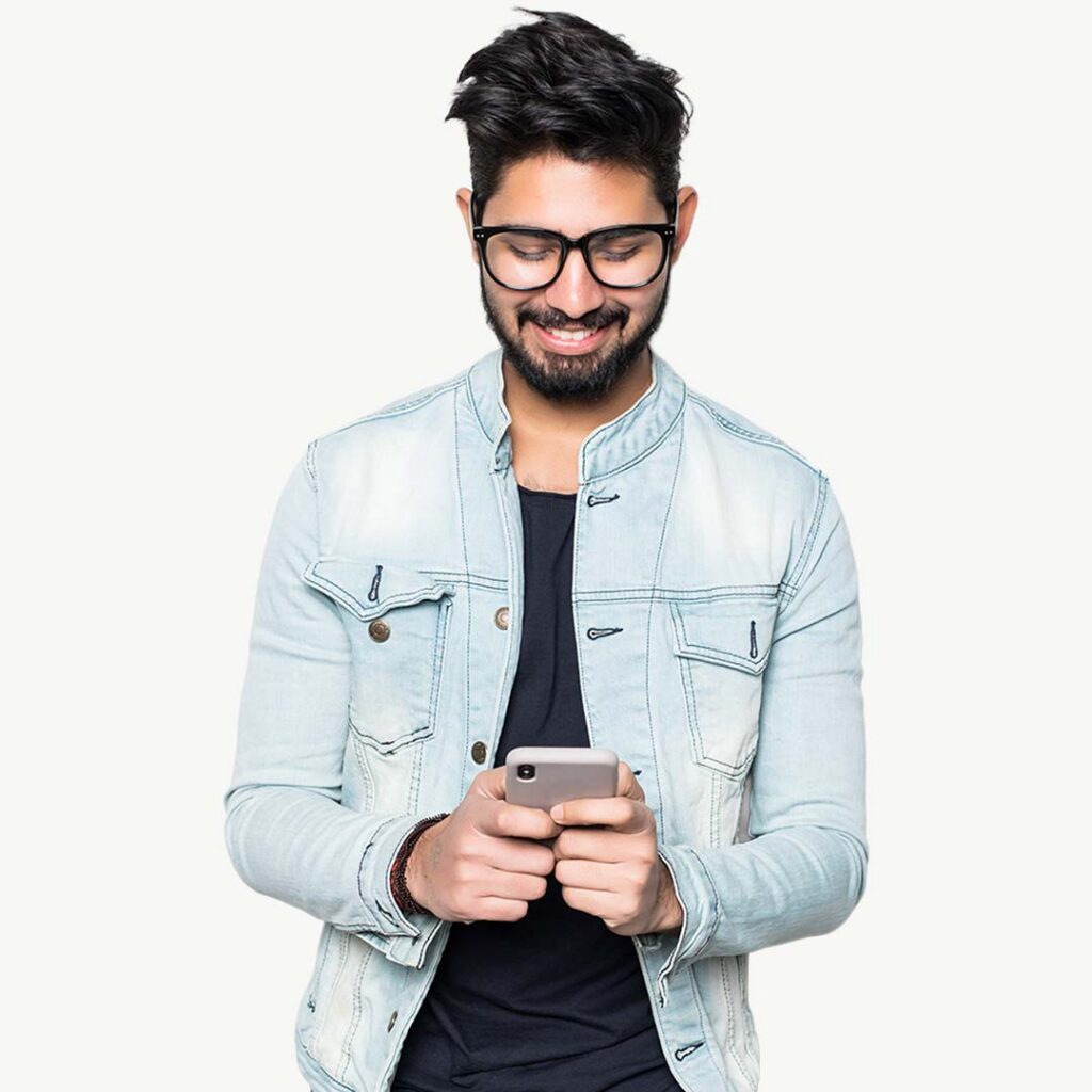 A smiling man wearing glasses and a light blue denim jacket over a black shirt, looking down at a smartphone he's holding against a white background.