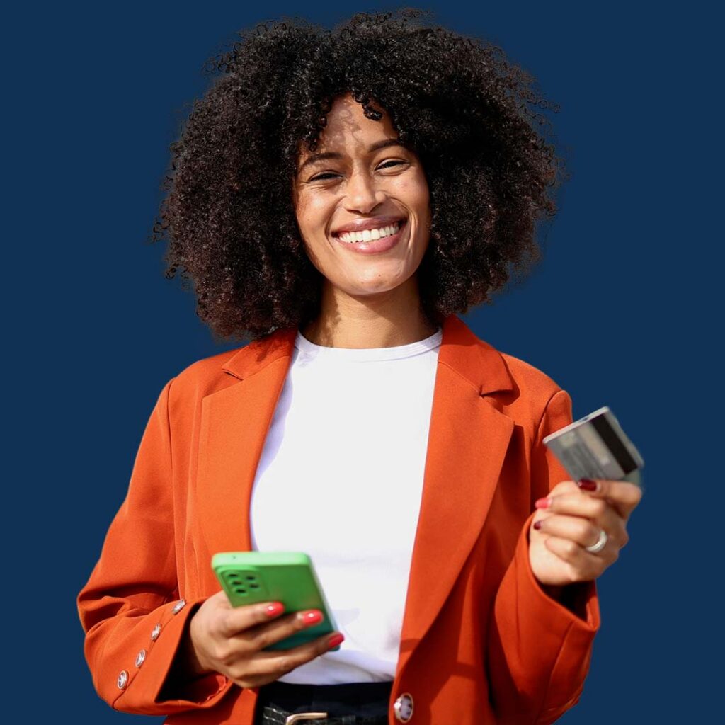 A smiling woman with curly dark hair wearing a bright orange blazer over a white shirt, holding a green smartphone in one hand and a gray credit card in the other, against a blue background.