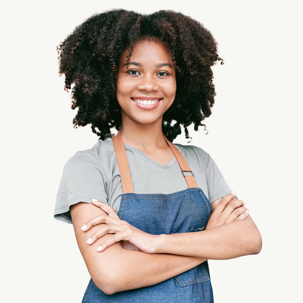 Portrait of a young woman with curly dark hair wearing a gray t-shirt and denim apron with tan straps, smiling at the camera with arms crossed against a white background.