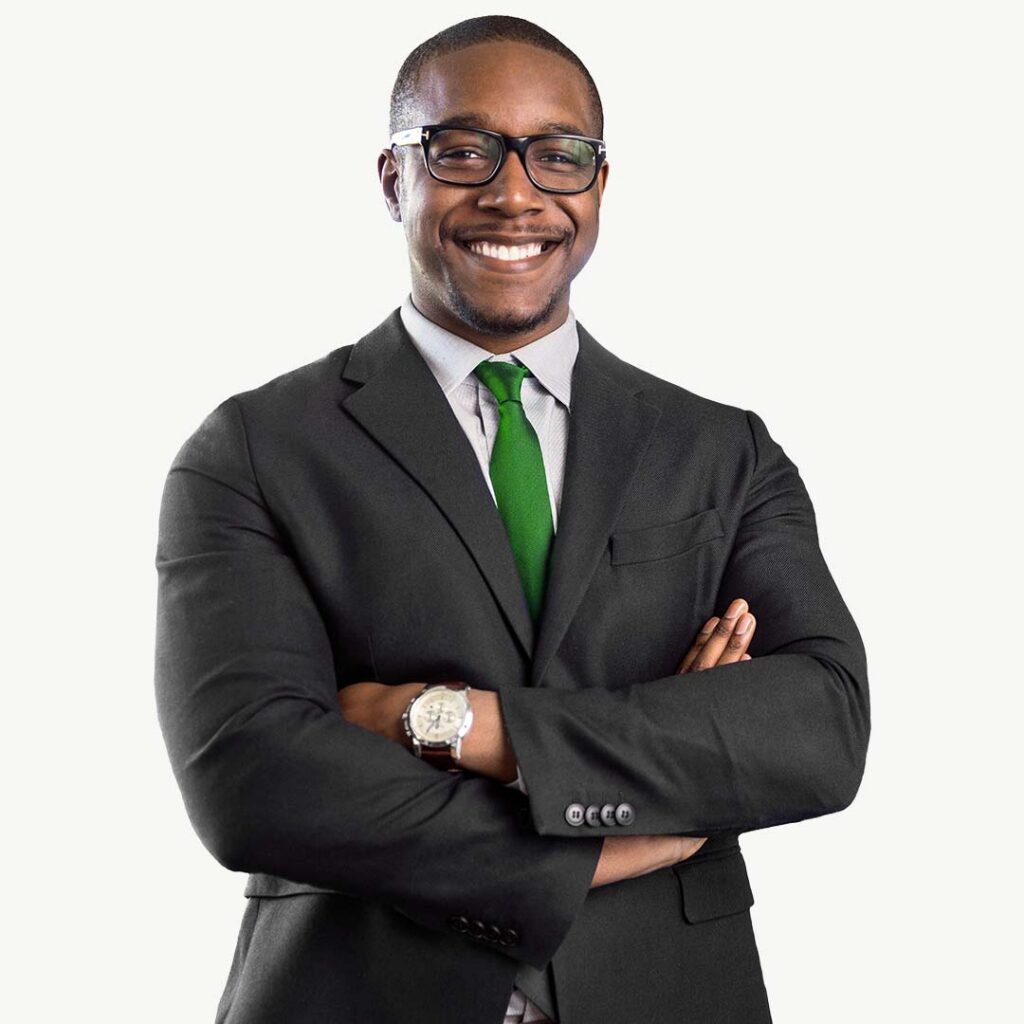 Professional headshot of a smiling man wearing a dark charcoal suit, light blue dress shirt, and green tie, with glasses and a watch, standing with arms crossed against a white background.