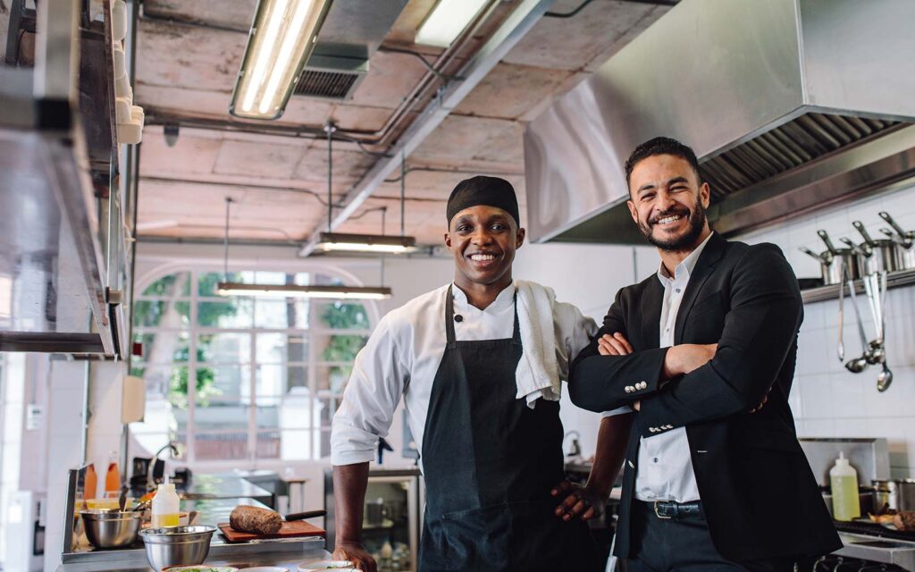 Two smiling men standing in a professional kitchen, one wearing a chef's apron and headwrap, the other in business attire with crossed arms, with cooking equipment and ingredients visible in the background.