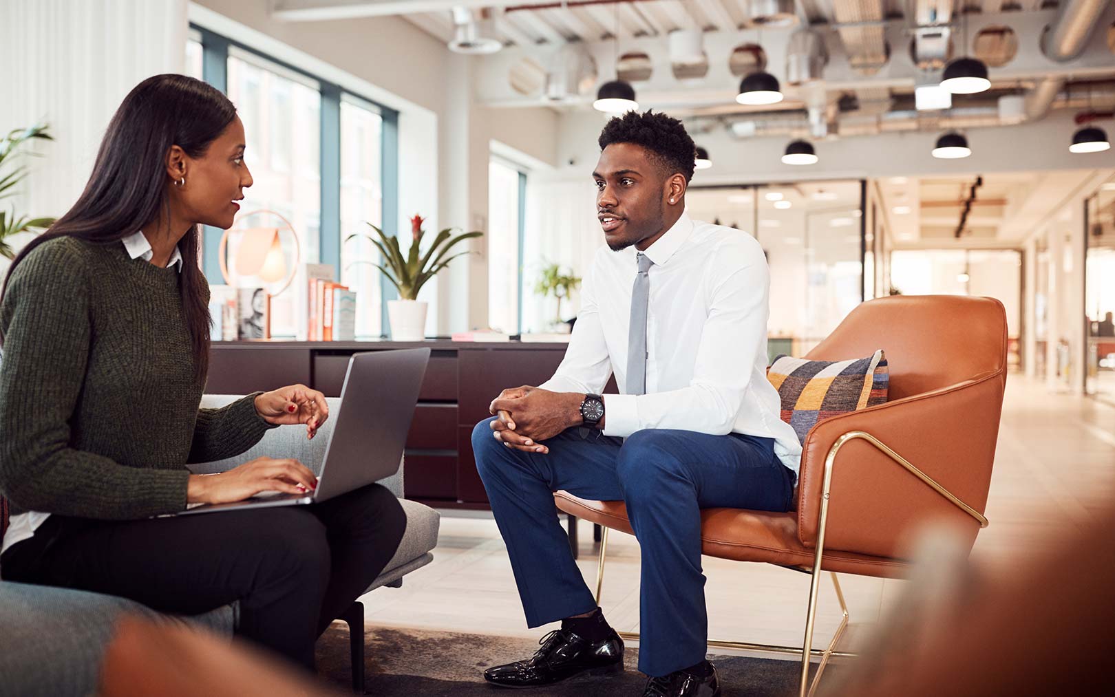 Two professionals having a conversation in a modern office lounge area. A woman on the left holds a laptop while wearing a dark green cardigan, and a man on the right sits in a tan leather chair wearing a white dress shirt and gray tie with dark blue pants. The bright, open office space features contemporary furniture, potted plants, and pendant lighting.