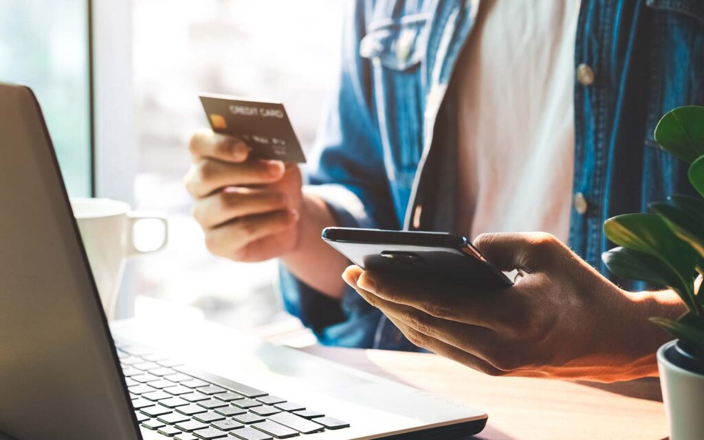 Person holding credit card and smartphone while working on laptop at desk with coffee cup and plant nearby.