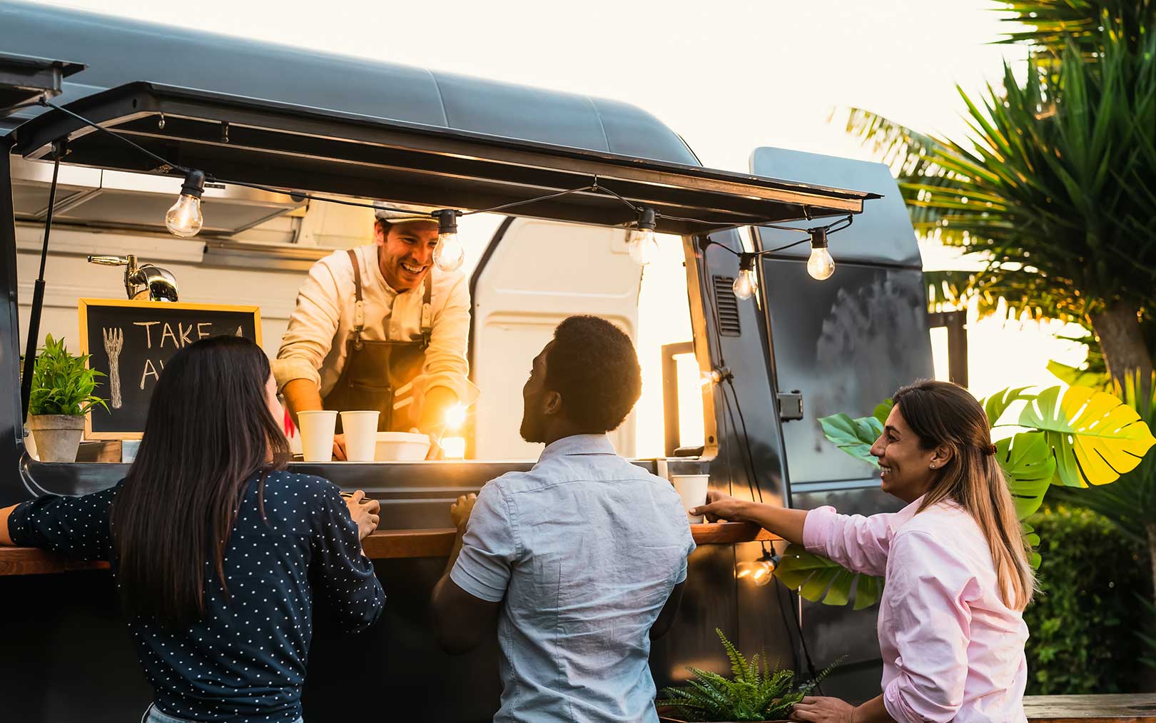 A smiling vendor serves food from a food truck window to three customers standing outside during evening hours, with string lights and tropical plants visible in the background.