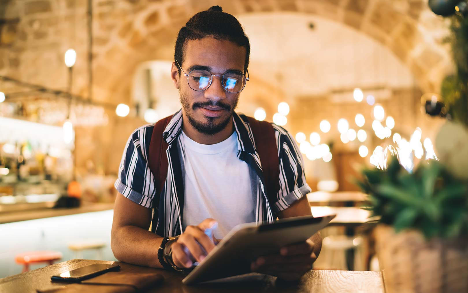 A man wearing glasses and a striped shirt works on a tablet at a wooden desk in a warmly-lit indoor space with architectural details and plants in the background.