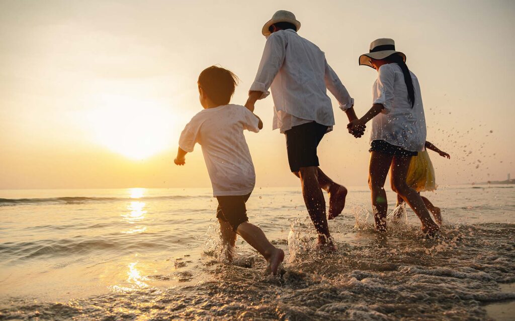 A family of four running into the ocean at sunset, with two adults wearing hats and light-colored shirts holding hands with two children in the shallow water.