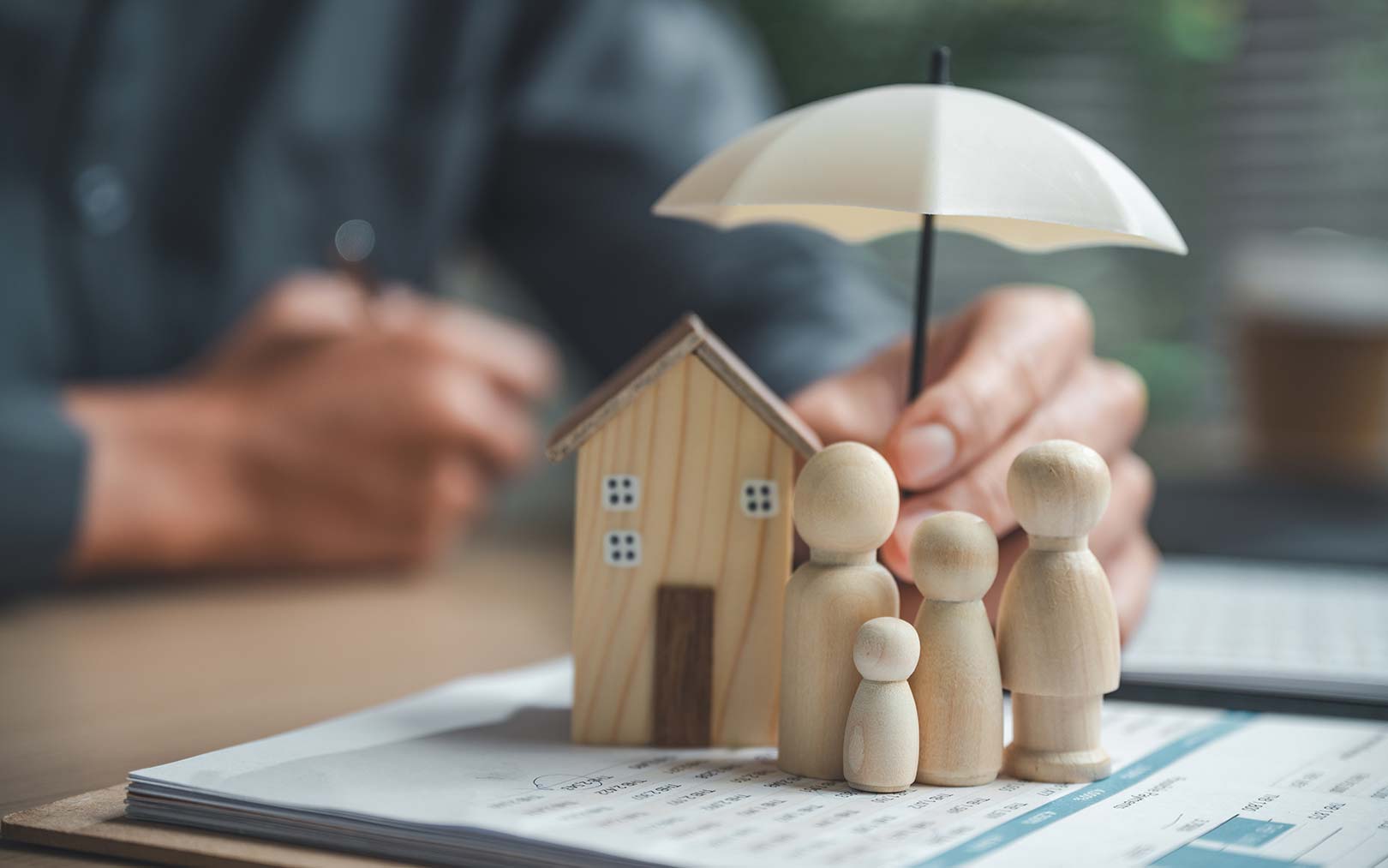 A person holds a small umbrella over wooden figurines representing a family standing next to a miniature house on financial documents, symbolizing home insurance and family protection.