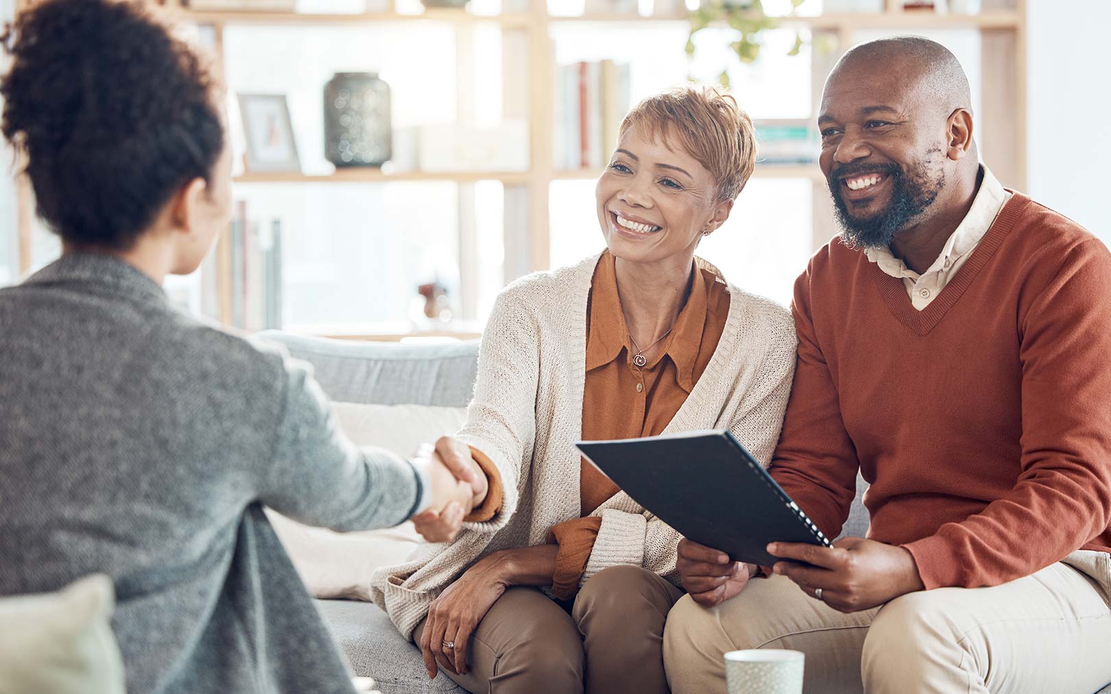 A professional couple shaking hands with a client during a consultation in a bright, modern office setting, with the woman holding a tablet while both smile warmly.