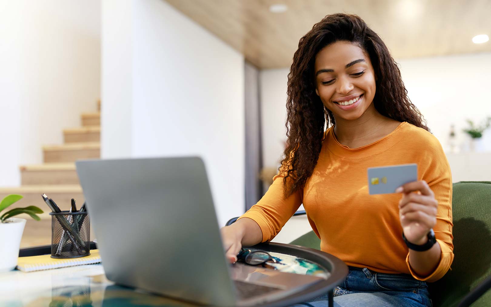 A smiling woman in an orange long-sleeve shirt sits at a desk with a laptop, holding a blue credit card while looking at the screen in a bright, modern office space.
