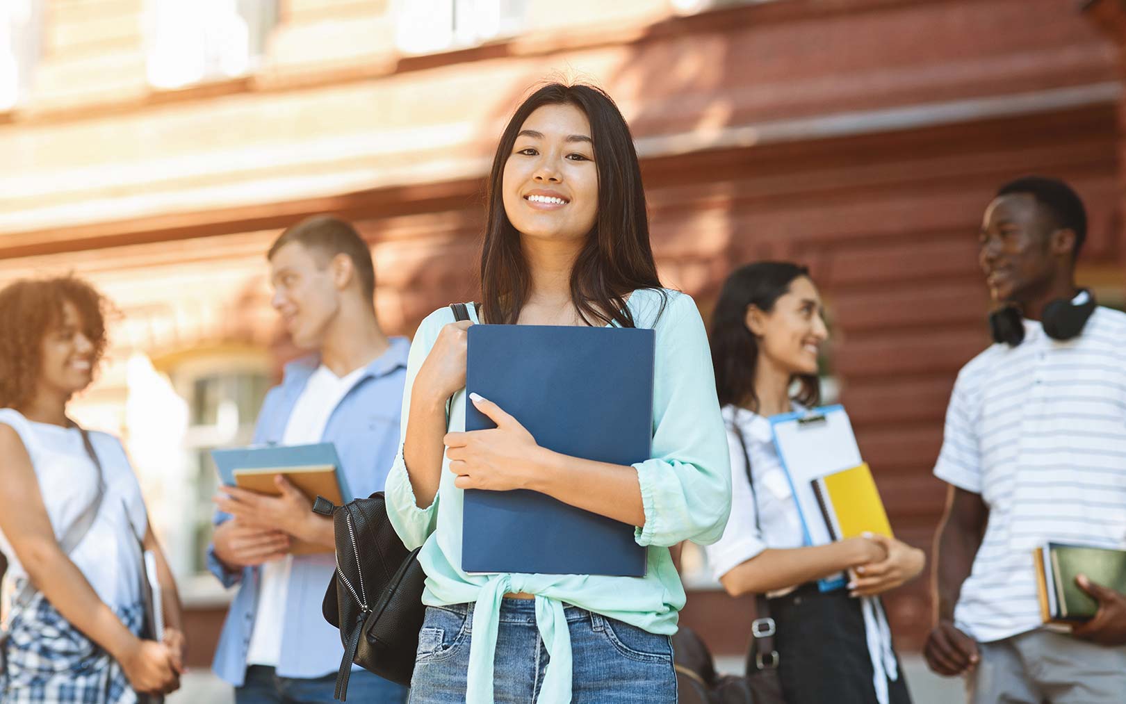A smiling young Asian woman holding a blue notebook and backpack stands in focus in front of a brick building, with diverse students holding books blurred in the background.