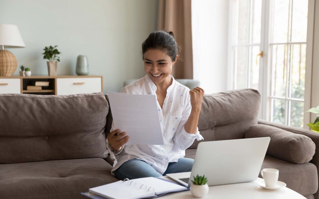 A woman sitting on a gray couch in a bright living room, smiling while holding papers and working on a laptop, with a coffee cup and small plant on the table beside her.