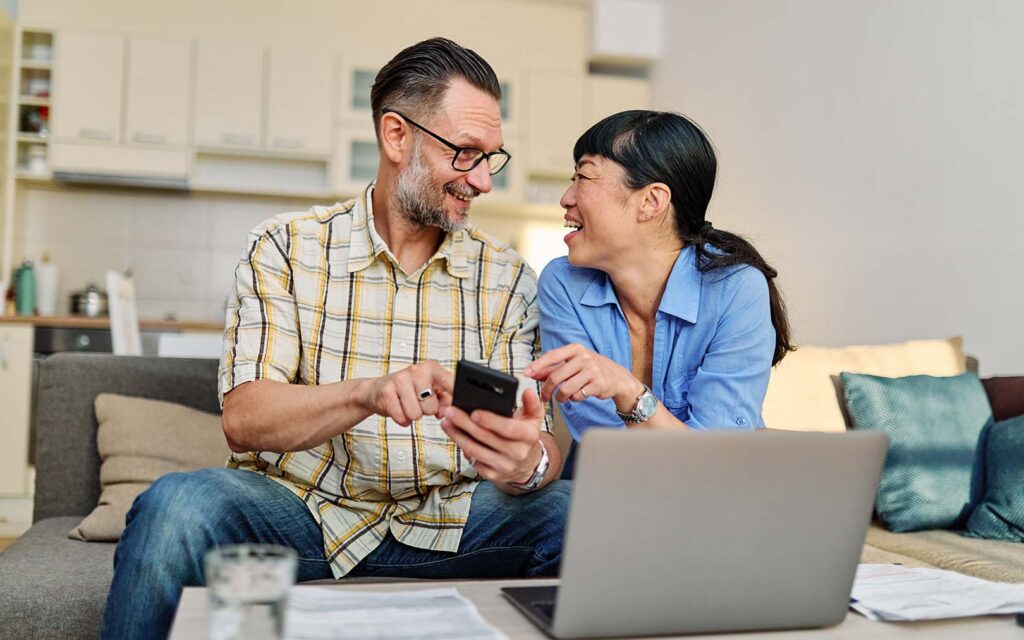 A man and woman sitting on a couch in a living room, smiling while looking at a smartphone together with a laptop and paperwork in front of them.