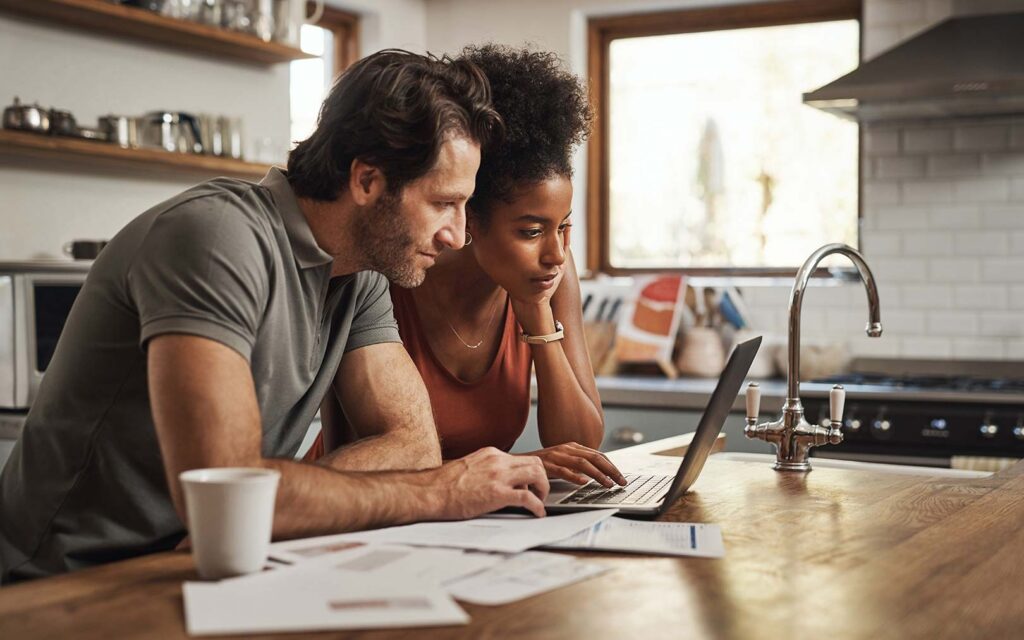Two people reviewing documents on a laptop at a kitchen counter, with papers and a coffee cup on the wooden surface.