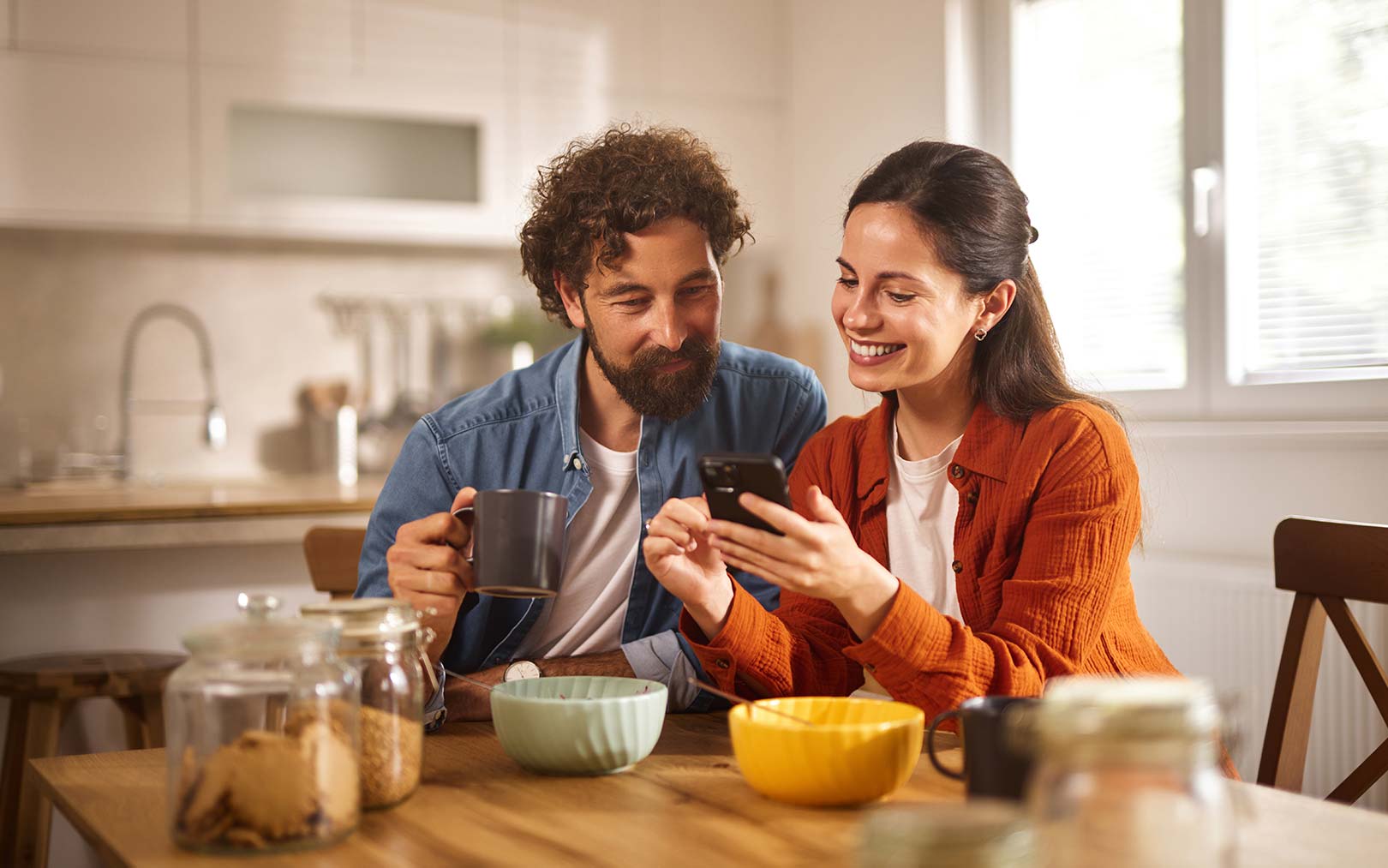 A smiling couple in a modern kitchen, with the woman showing the man something on her smartphone while holding a mug of coffee. They are seated at a wooden table with bowls and kitchen items visible.