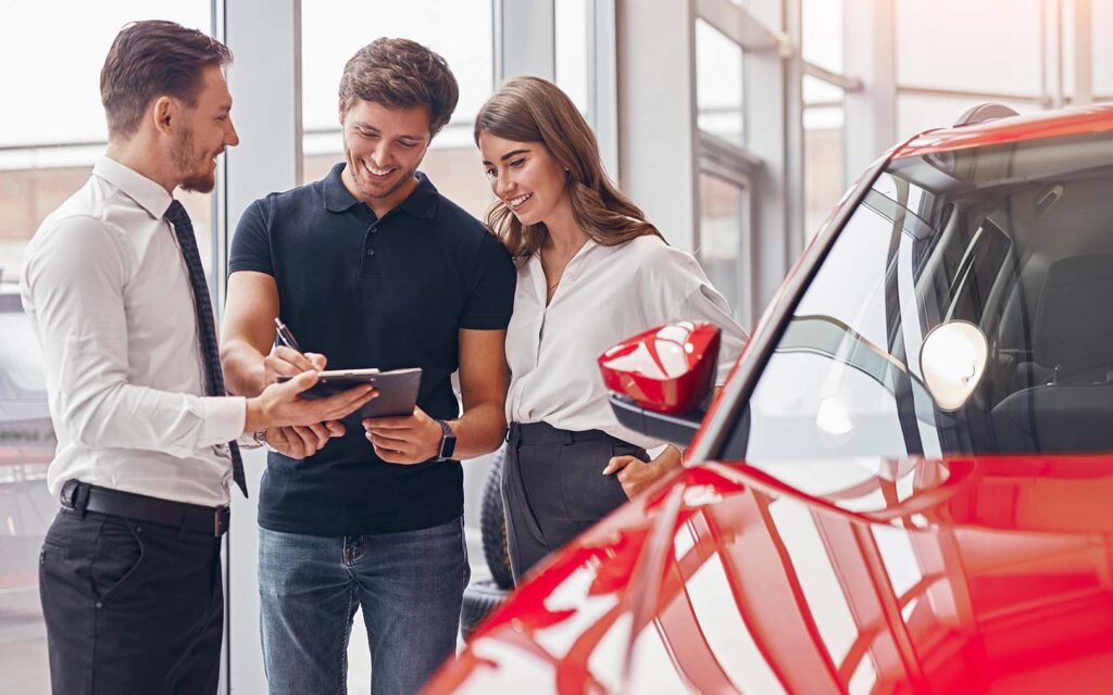 A car salesman in formal attire showing a red vehicle to a young couple in a modern dealership showroom, with the salesman holding a tablet while discussing the car.