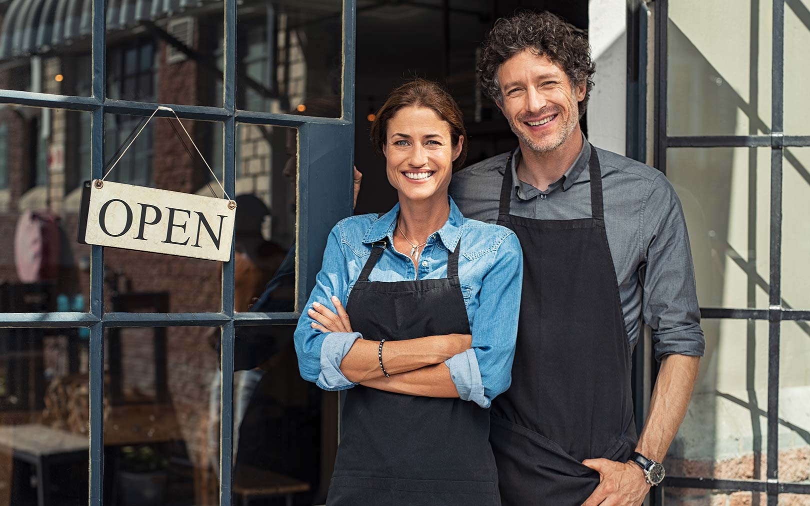 Two smiling business owners standing in the doorway of their shop, wearing black aprons, with an "OPEN" sign hanging on the glass door behind them.