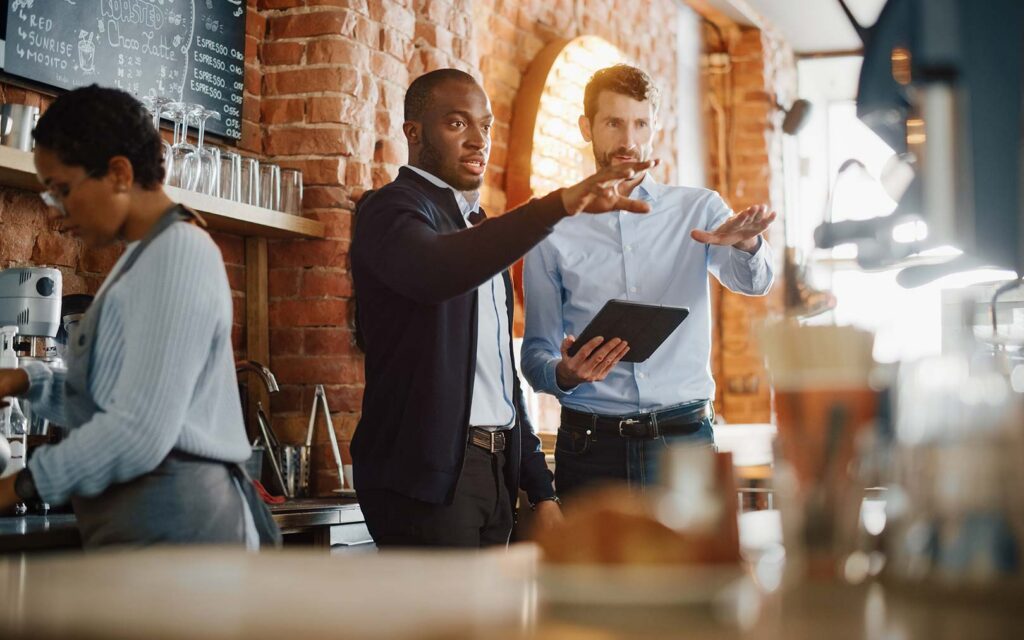 Two businessmen discussing work at a coffee shop counter, one holding a tablet while gesturing, with a barista in the background and exposed brick walls visible.