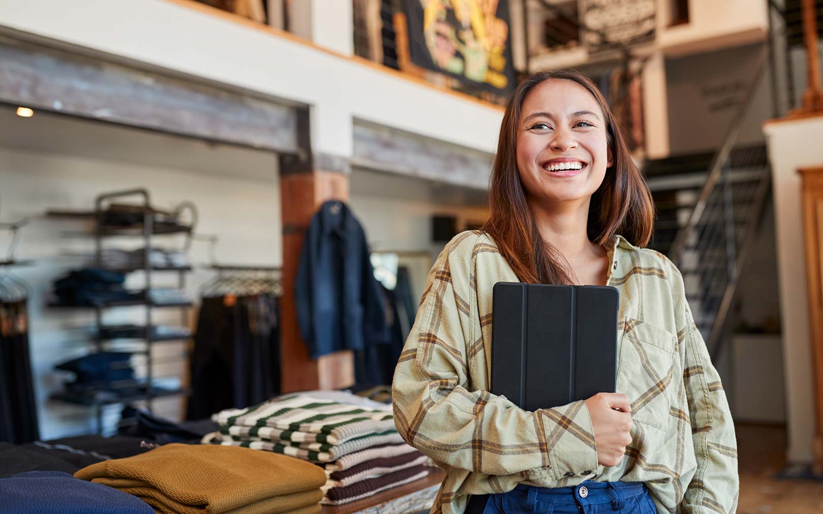 A smiling young woman holding a tablet stands in a clothing retail store, wearing a plaid shirt, with folded clothes and shelving visible in the background.