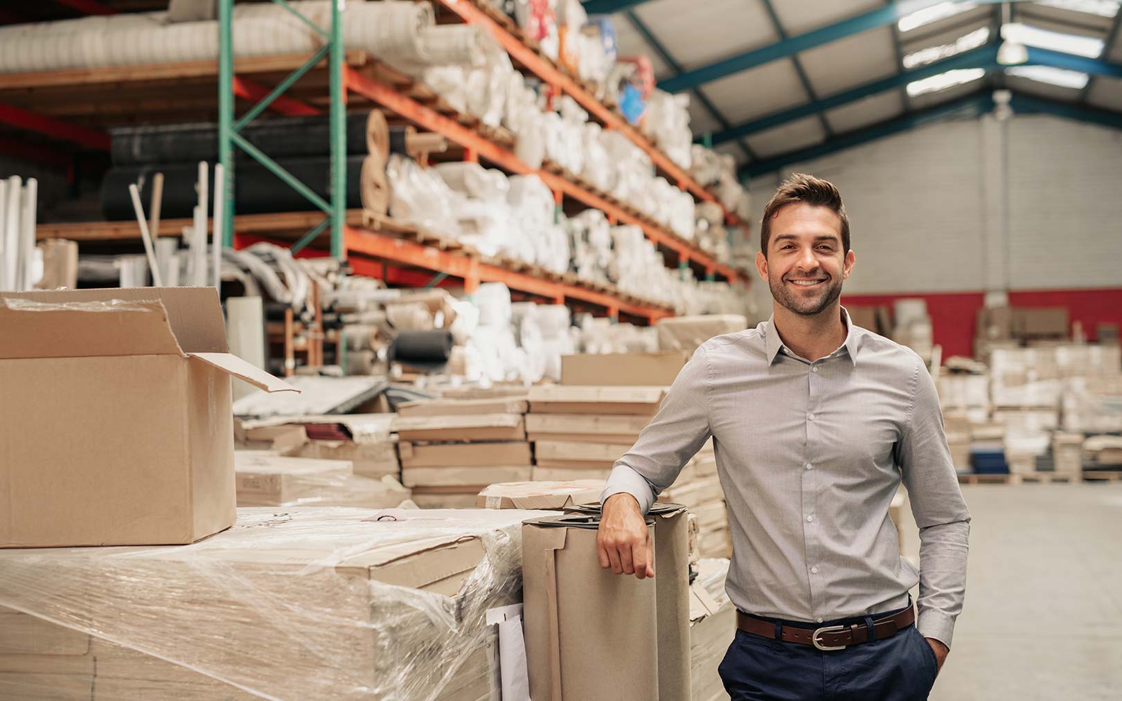 A smiling man in a light gray shirt and dark pants stands in a large warehouse filled with cardboard boxes, wooden materials, and industrial shelving with orange and green racks.