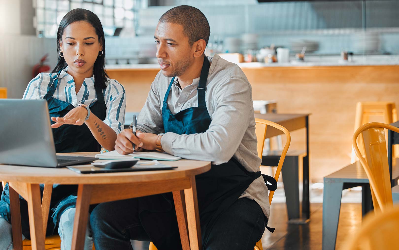 Two restaurant workers in aprons review documents and a laptop together at a wooden table in a modern kitchen.