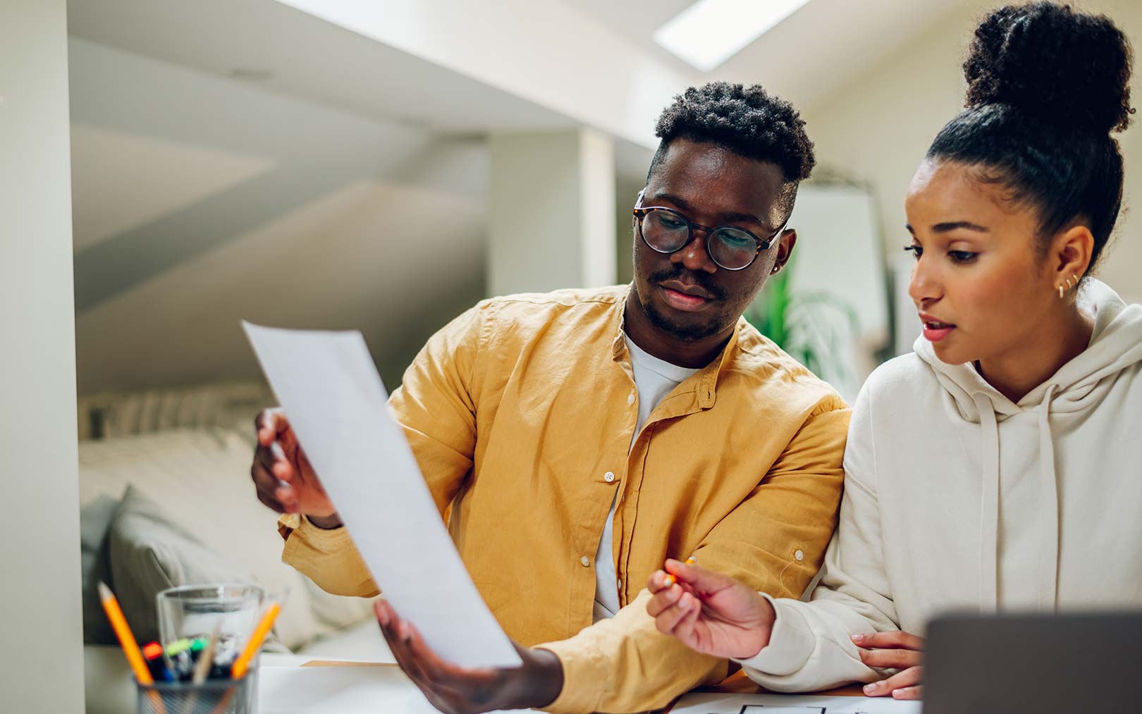 Two young professionals collaborating at a desk, with a man in a yellow shirt holding a tablet while discussing with a woman in a cream hoodie in a modern office environment.