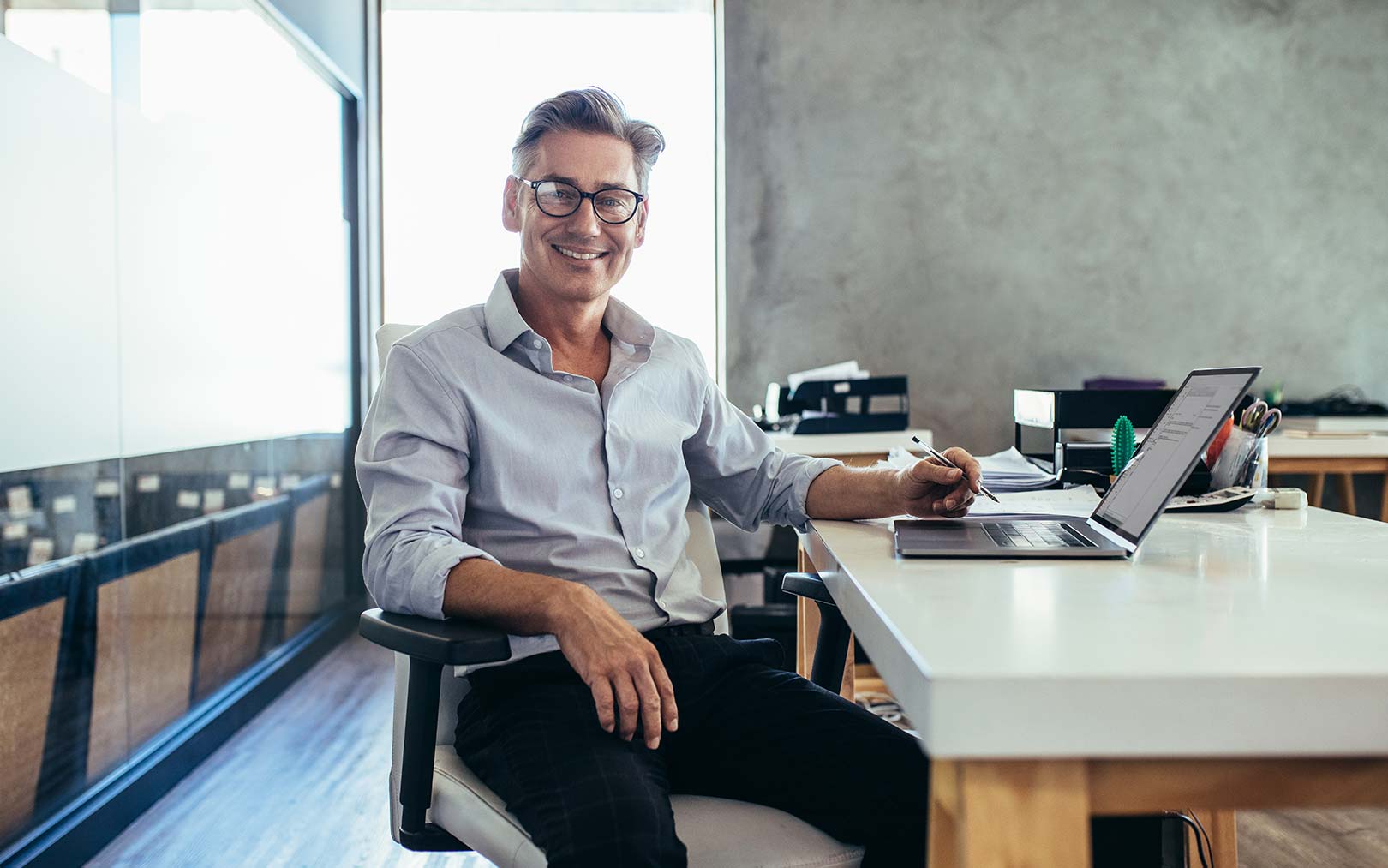 A smiling man wearing glasses and a light blue button-up shirt sits at a desk in a modern office, holding a stylus while working on a tablet, with a laptop and office supplies nearby.