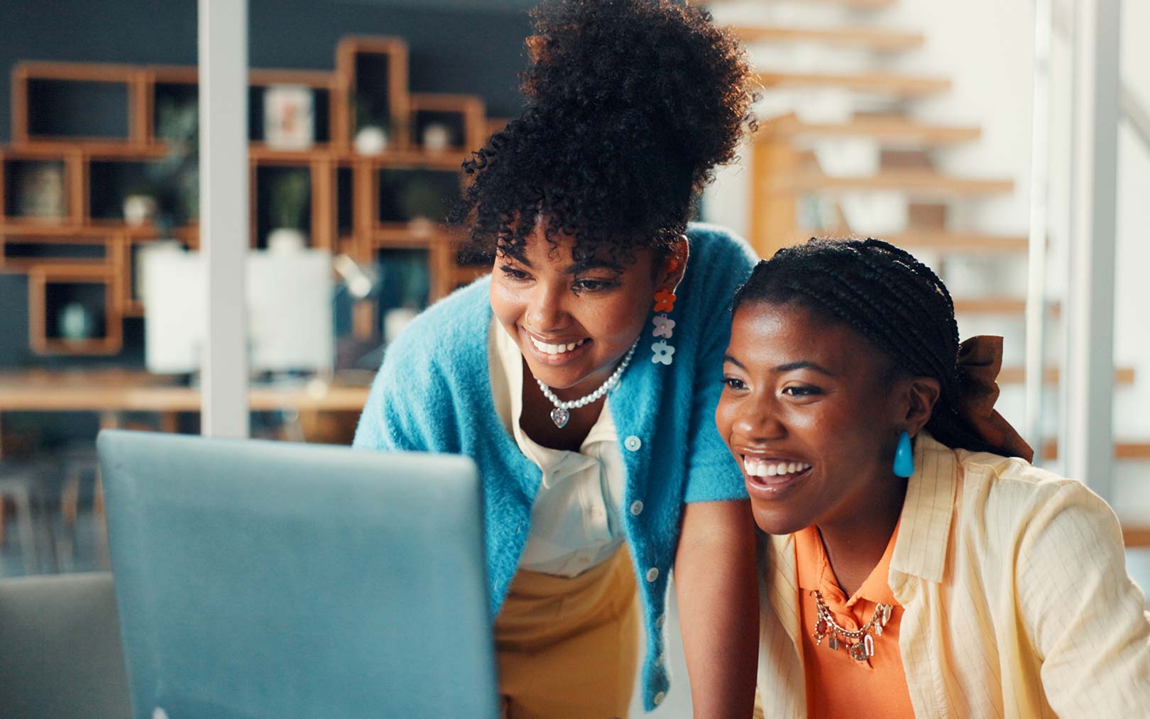 Two smiling women looking at a laptop computer together in a bright, modern office space with wooden shelving in the background.