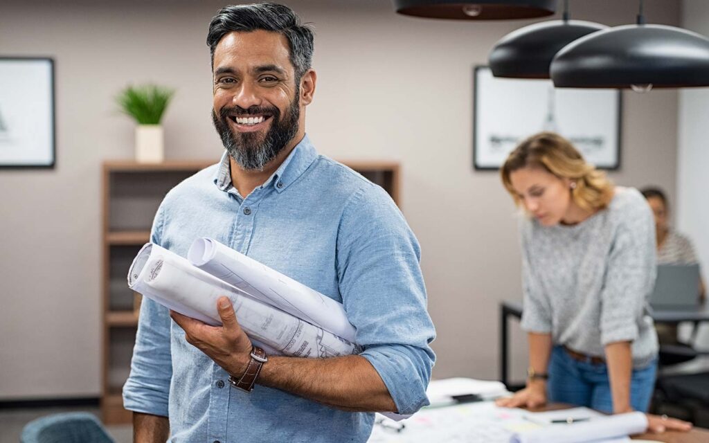 A smiling man with a beard wearing a light blue shirt holds rolled blueprints in a modern office, with two colleagues working at a desk in the background.