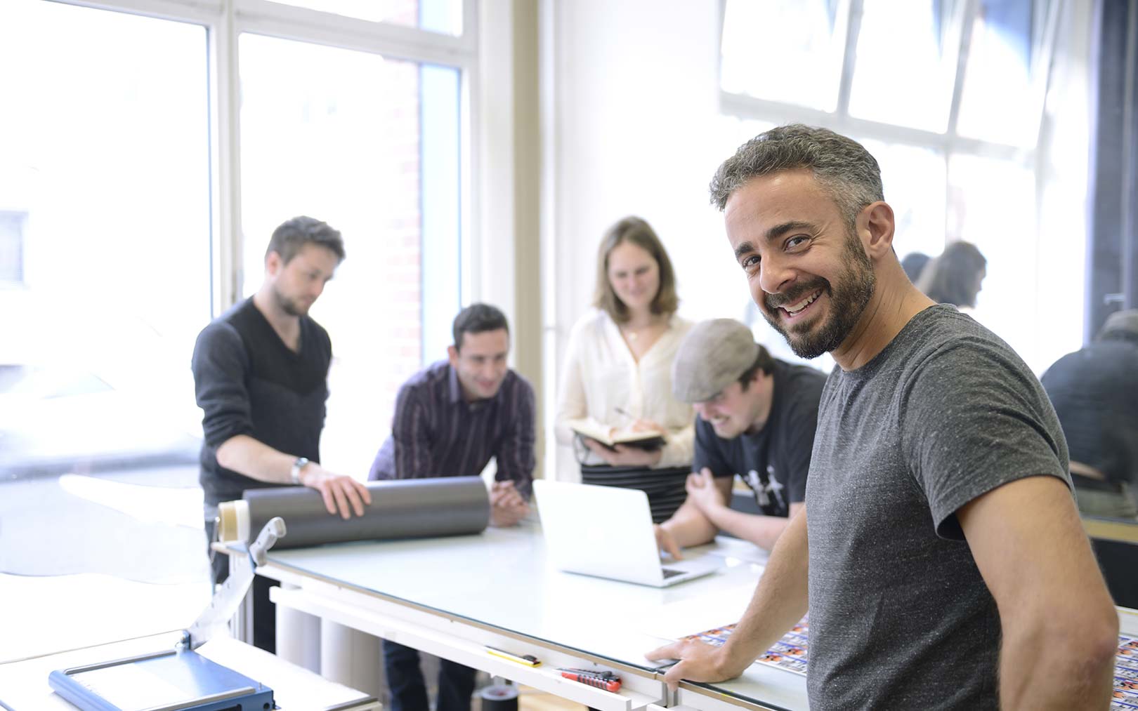 A man in a gray t-shirt smiles at the camera while working at a table in a bright, modern classroom or workshop space with large windows. Several other people are visible in the background engaged in collaborative work with laptops and materials.