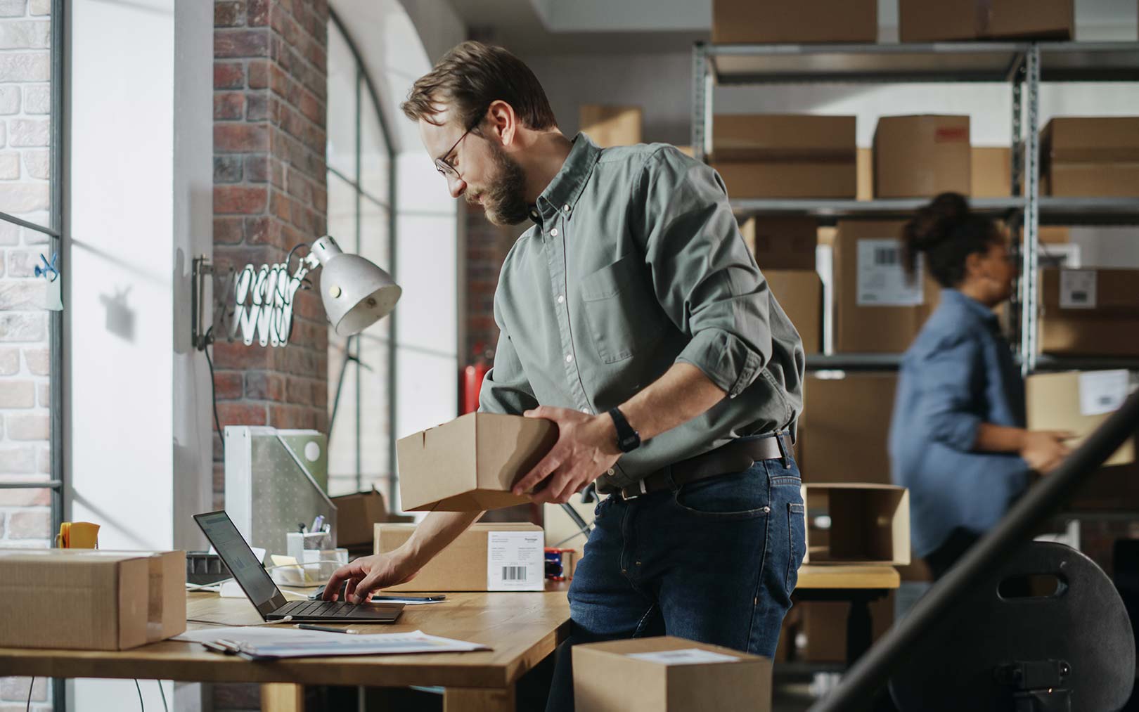 A man wearing glasses and a gray shirt holds a cardboard box while working at a desk with a laptop in a warehouse or fulfillment center, with another person visible in the background near shelving units.