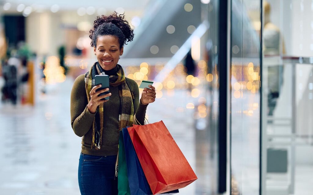 A smiling woman shopping in a mall holds a smartphone and credit card while carrying colorful shopping bags past a storefront with blurred bokeh lights in the background.