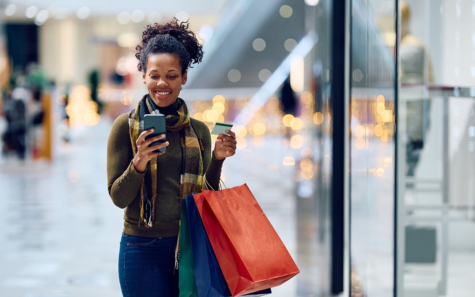 A smiling woman shopping in a mall holds a smartphone and credit card while carrying colorful shopping bags past a storefront with blurred bokeh lights in the background.