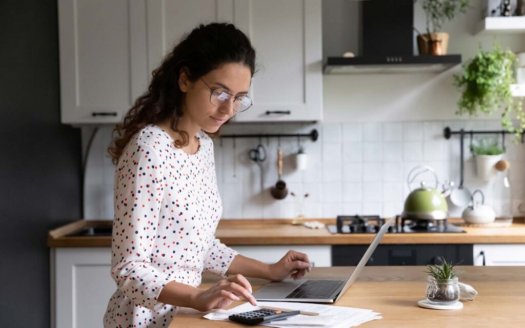 A woman wearing glasses and a polka dot blouse works on a laptop at a wooden kitchen counter, with a calculator and documents nearby, in a modern kitchen with white cabinets and stainless steel appliances.