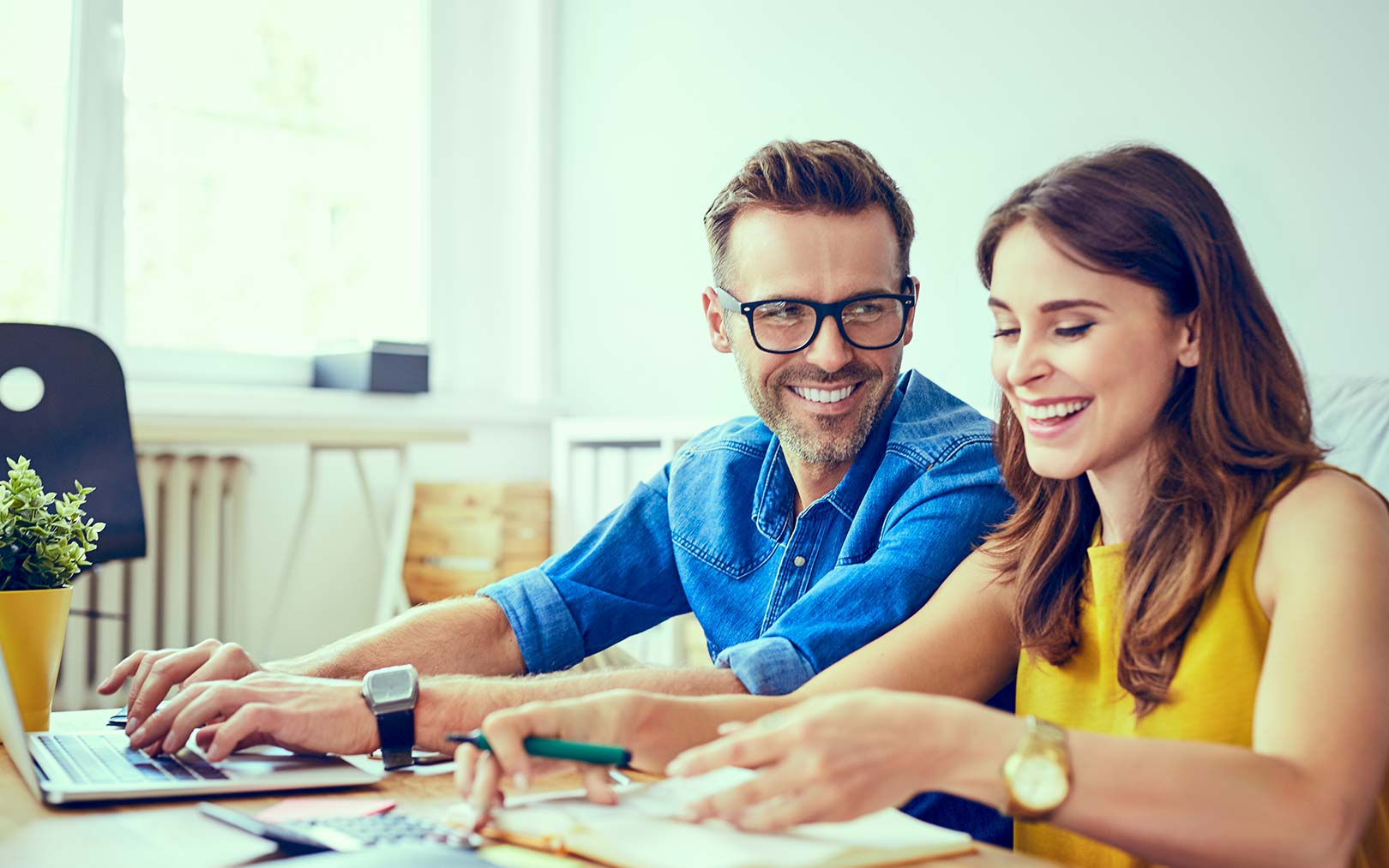Two smiling professionals collaborating at a desk with a laptop, documents, and office supplies in a bright, modern workspace.