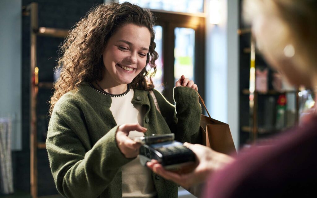 A smiling woman with curly brown hair wearing a green cardigan and white shirt holds a wallet while making a contactless payment with a card reader held by another person in what appears to be a retail setting.