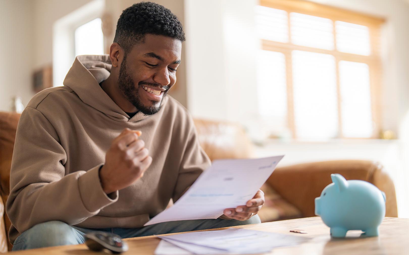 A smiling man in a beige hoodie sits at a wooden table reviewing documents, with a blue piggy bank and papers visible, while sunlight streams through windows in the background.