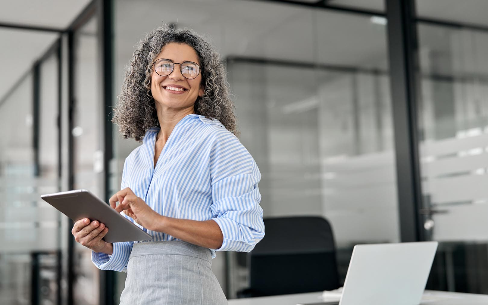 Professional woman with curly gray hair wearing glasses and a blue striped shirt, holding a tablet in a modern office environment with glass partitions and a laptop visible on the desk.