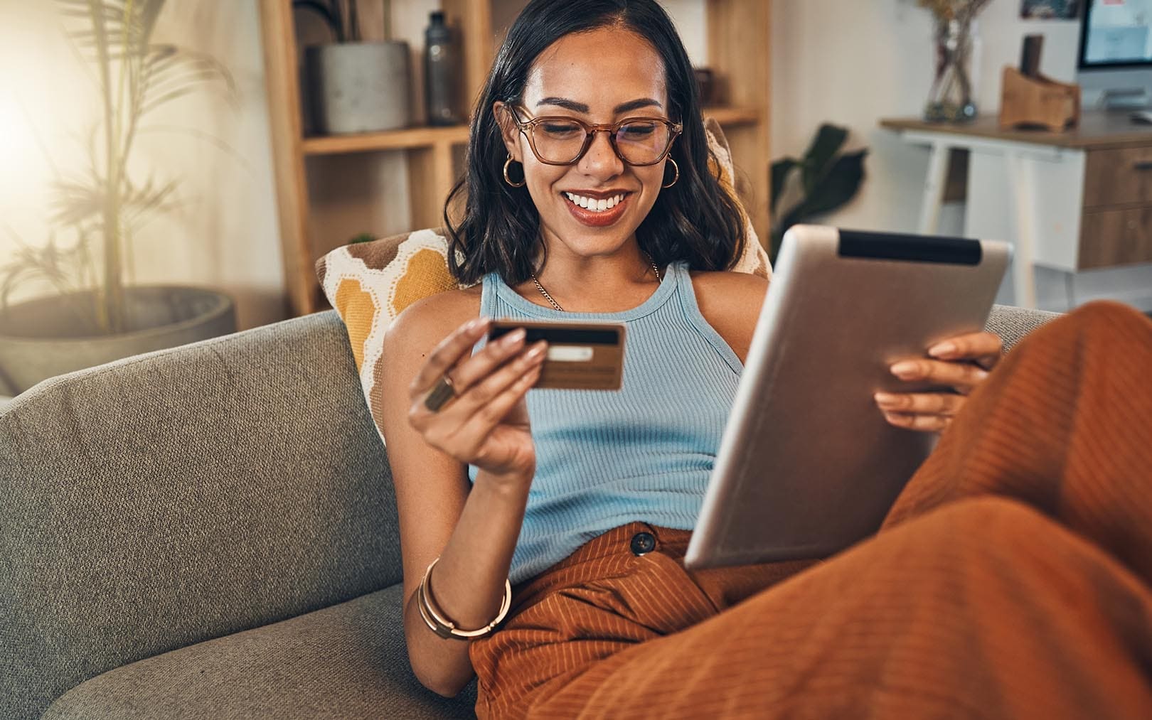 Woman smiling while shopping online on a tablet and holding a credit card on a couch at home.