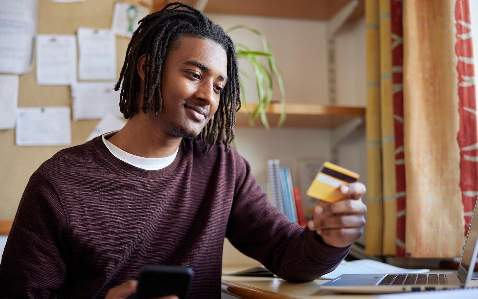A young man with dreadlocks wearing a maroon sweater holds a yellow credit card while looking at his smartphone and laptop in an office setting with a cork bulletin board and plant visible in the background.