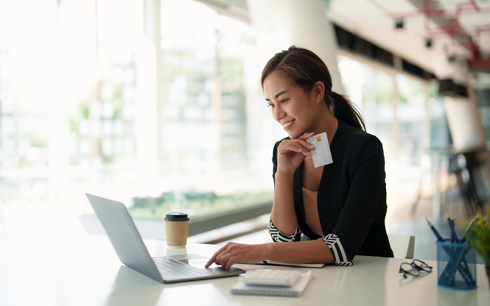 A smiling woman in a black blazer sits at a desk with a laptop and coffee cup, holding a credit card while working in a bright, modern office space with large windows.