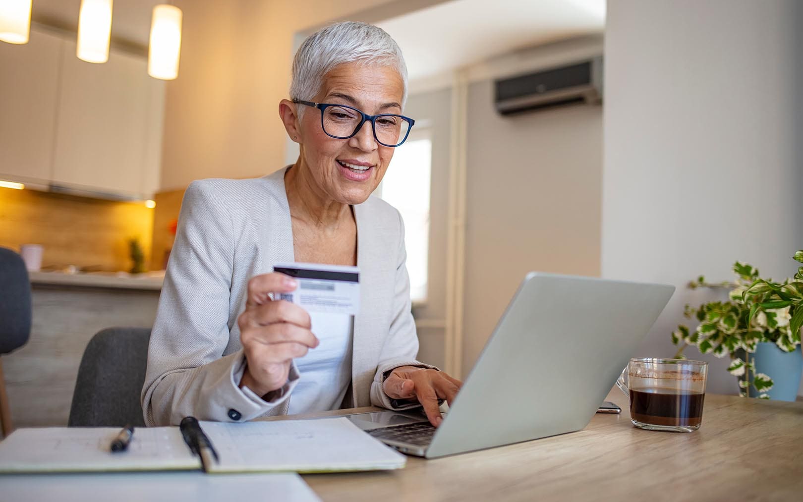 Smiling senior woman wearing glasses and beige cardigan holding a credit card while using a laptop at a desk in a modern kitchen.