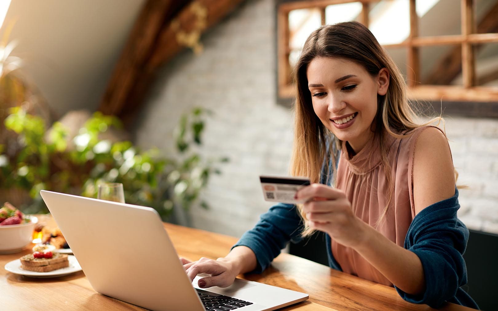 A woman smiling while holding a credit card and using a laptop at a wooden table in a bright, plant-filled room, appearing to make an online purchase.