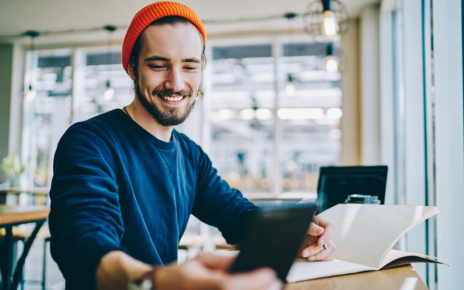 A smiling man wearing an orange beanie and dark blue shirt sits at a table by large windows, holding a tablet and looking at an open notebook in a bright, modern office space.