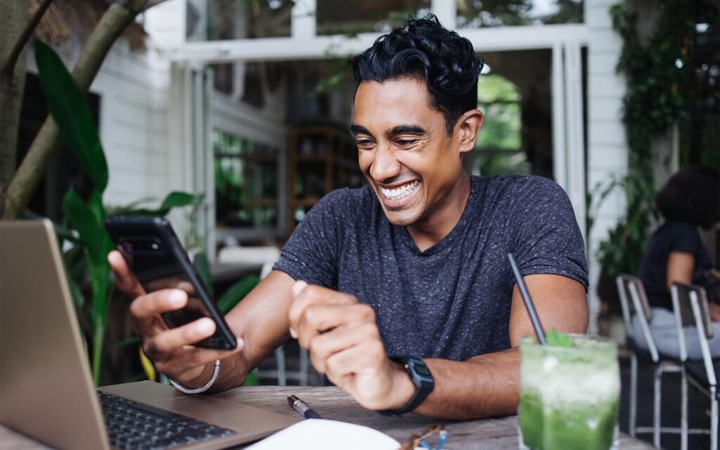 A man in a dark gray t-shirt smiling while holding a smartphone at an outdoor table, with a laptop and beverage nearby, and a white house with windows visible in the background.
