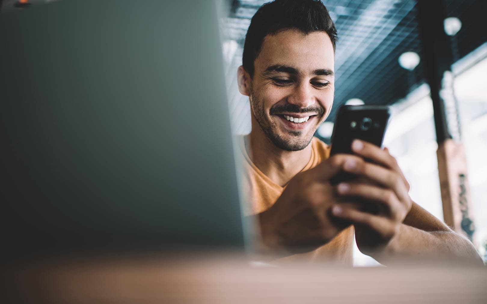A smiling man in a yellow shirt looking at his smartphone in an indoor setting with modern lighting.