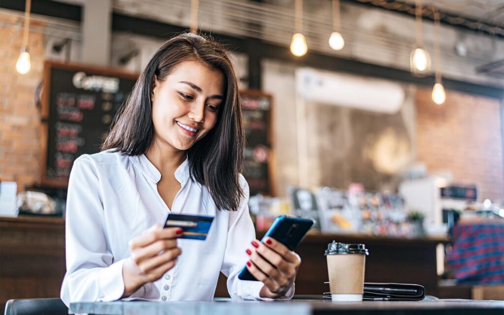 A smiling woman in a white blouse holds a credit card and smartphone while sitting at a table in a modern café, with a coffee cup beside her.